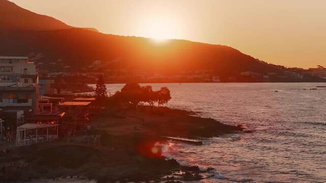 A serene sunset in Stalida, Crete, with mountains, calm waters, rocky shoreline, boats, lit terraces, and people strolling along the coast.
