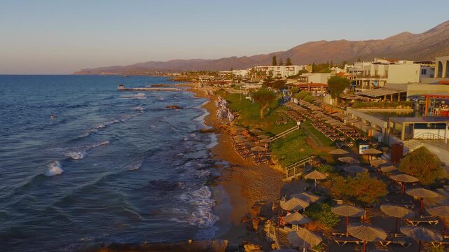 Stalida's beachfront at sunset with blue and white umbrellas, gentle waves, a small pier, whitewashed buildings, and mountains in the distance.
