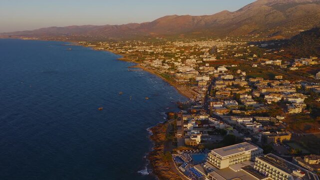 A sweeping aerial view of Stalida, Crete, showing sandy beaches, calm waters with boats, white buildings, and golden lit mountains during sunset.