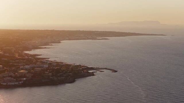 View of Stalida on Crete during sunset, with white buildings, calm sea, rugged hills, and smooth camera motion capturing the tranquil coastline.
