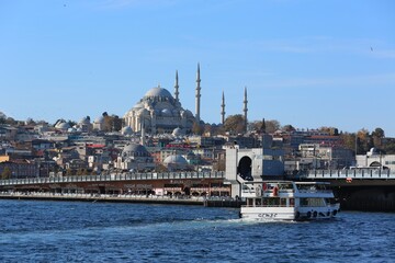 Obraz premium A scenic view of the Süleymaniye Mosque rising above Istanbul’s skyline, with the Galata Bridge and a ferryboat crossing the Golden Horn under a bright blue sky.