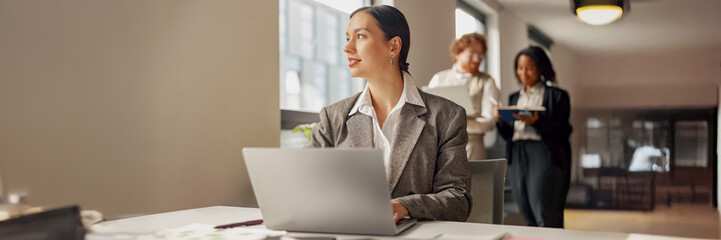 A dedicated professional works on her laptop while team members collaborate in a modern, innovative workspace
