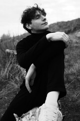 Fototapeta premium Emotion black and white portrait of a young guy in a black wear. Man have relax and smile. Seating on the tree