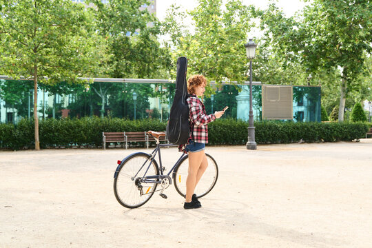 Young musician checking her phone in the park with bicycle and guitar - Powered by Adobe