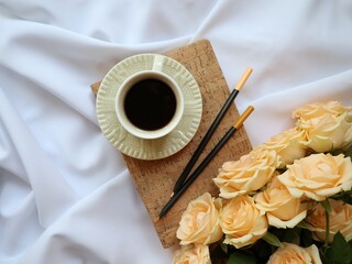 Cup of black coffee with peach roses and pencils on cork board over white fabric. Cozy morning flat lay.