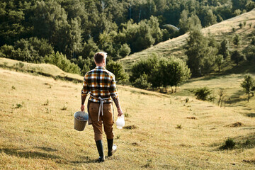 Modern farmer strolling through sunlit countryside carrying milk and a bucket