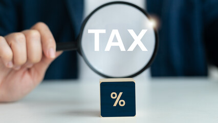 Close-up of a magnifying glass focusing on the word TAX with a percentage block, symbolizing tax...