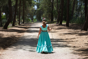 Fototapeta premium A beautiful young Spanish woman dressed in a traditional green flamenco dress with a flower in her hair walks among the pine trees at the pilgrimage, leaning on a stick. Photo taken from behind.