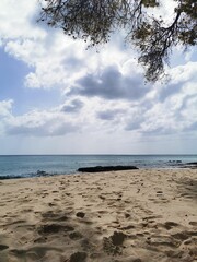 A wide, serene view of a sandy tropical beach under a dramatic cloudy sky, with calm turquoise waters and a dark rock formation