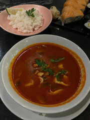 A close-up of a Japanese meal including spicy soup, white rice with lemon, and assorted sushi rolls on a black table.