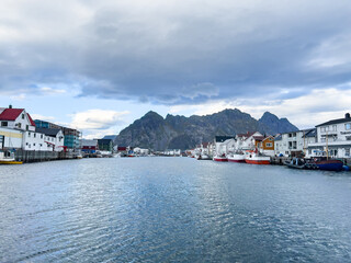 Naklejka premium A scenic view of Henningsvær in Lofoten Islands, northern Norway, showcasing traditional buildings along the harbor with mountains in the background.