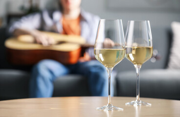 Glasses with white wine on table and man playing guitar indoors, selective focus