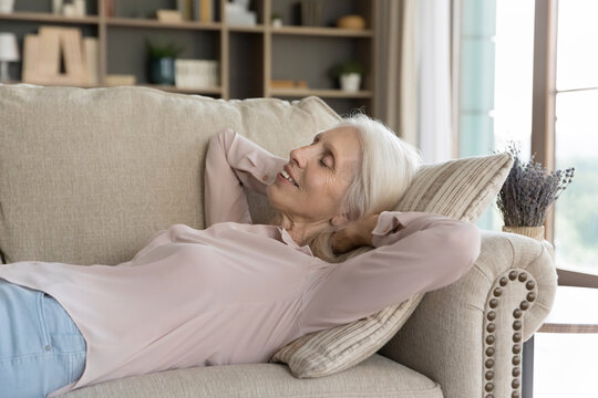 Carefree daytime nap. Smiling tranquil senior adult lady pensioner lying on comfy sofa in relaxed pose dreaming sleeping on back with hands behind head breathing deep enjoy quiet moment of serene rest