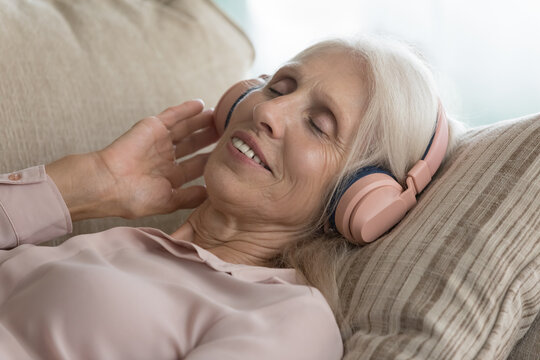 Digital me time. Close up portrait serene elderly female retiree in modern wireless headphones relaxing on cozy sofa with closed eyes listen to calm music from radio or online playlist enjoy audiobook