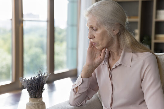 Grieving in silence. Weak upset lonely elderly lady rest on couch by window lean head on hand suffer of aging problems solitude isolation think about death bad health in sad depressed mood. Copy space