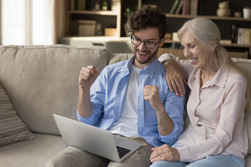 Fototapeta premium We did it. Overjoyed adult son hold notebook on lap raise hands up in victory gesture celebrate online success together with senior mother winning internet game getting bonus lottery prize. Copy space