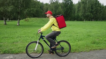 A delivery man takes a food order using a smartphone and rides a bicycle with a red food delivery bag on his shoulders along the road against the background of a city park