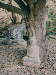 Obraz premium Serene Buddha statue beside a flowing stream under a tree in a tranquil garden setting