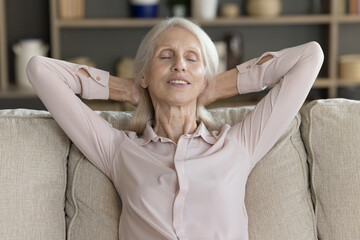 Unwinding with comfort. Happy smiling older age woman on retirement lean back on soft cozy couch relax holding hands behind head feel peace of mind sleepy enjoy light carefree nap dream at home alone