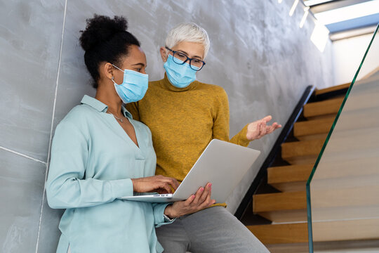 Women in startup workplace wearing masks, collaborating with laptop on creative projects in office