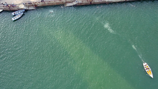 Aerial top-down view of the Capibaribe River in the Historic Center of Recife, Pernambuco. People swim and jump from the pier, with small boats docked and another navigating the green waters.