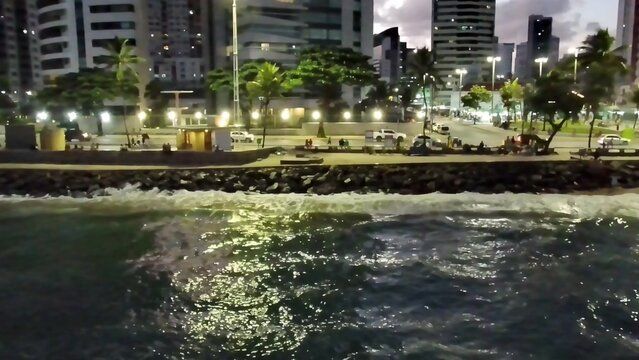 Night aerial view of the illuminated city skyline and waterfront of Boa Viagem Beach in Recife, Pernambuco, Brazil. Waves crash on the coast as traffic moves along the urban promenade.