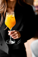 Close-up of a hand holding a wine glass with bright orange cocktail, ice cubes, and seeds. Blurred background. Stylish and refreshing summer drink concept