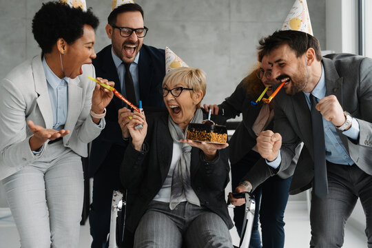 Group of business colleagues celebrating in a office with a birthday cake, party hats and blowers. The coworkers enjoying a fun in office party - Powered by Adobe