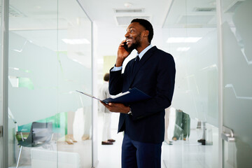 Businessman smiling while talking on the phone and holding documents in modern office corridor