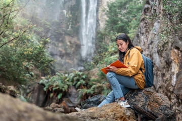 An Indonesian woman in a yellow jacket with a backpack sits on rocks near a waterfall, holding a notebook and pen, journaling in nature. Concept of mindfulness, travel, and inspiration.