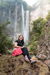 Obraz premium Smiling Asian woman sitting on rock with waterfall in background at Indonesia rainforest. Travel and adventure lifestyle concept showing joy, freedom, and connection with tropical nature.