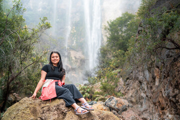Naklejka premium Indonesian woman sitting on a rock with a large tropical waterfall in the background. Concept of adventure, relaxation, eco tourism, freedom, nature travel, and exploration in lush rainforest.