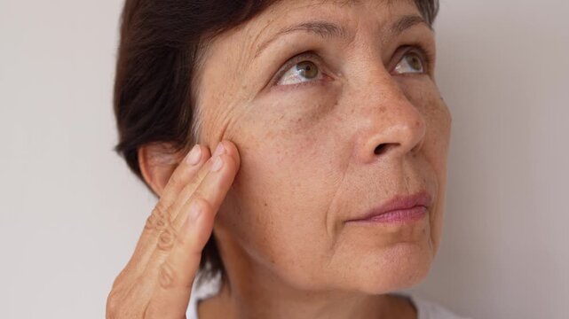 Close-up of mature woman gently touching the skin under her eye, showing wrinkles and puffiness on a light background. Visible signs of aging, natural beauty, slow motion