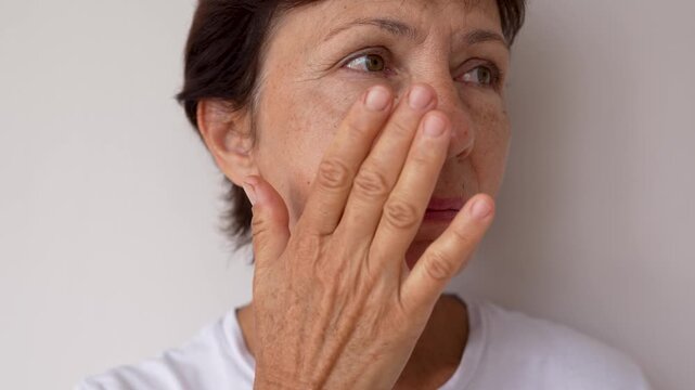 Close-up of mature woman with under-eye puffiness, wrinkles and age spots gently touching her face on a light grey background. Natural signs of aging, self-awareness and authentic beauty