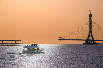 Obraz premium A boat navigates the Tamsui River against the backdrop of the iconic Tamsui River Bridge under construction in New Taipei City, Taiwan, at sunset. The sky is bathed in warm, soft hues.