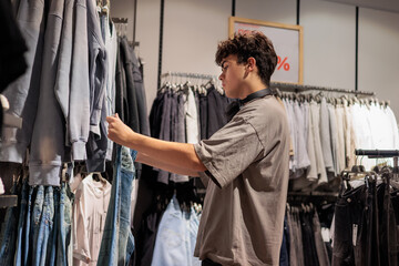 Young Man Choosing Jeans in a Clothing Store. Modern Fashion and Style.  Teen boy 16 years old shopping in apparel store, looking at jeans and denim clothes, modern teenager fashion lifestyle. 