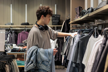 Teen boy 16 years old shopping in apparel store, looking at jeans and denim clothes, modern teenager fashion lifestyle. Adolescent boy in clothing shop choosing casual wear, youth style.
