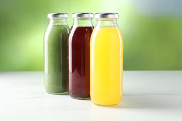 Tasty juices in glass bottles on white marble table against blurred green background, closeup