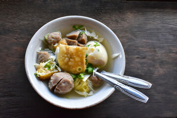 Meatball soup mixed with vegetables, noodles, egg in a bowl with cutlery on a wooden board. Indonesian hearty soup served at the market.