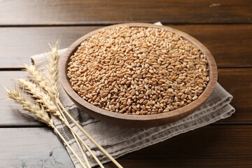Wheat grains in bowl and spikelets on wooden table, closeup