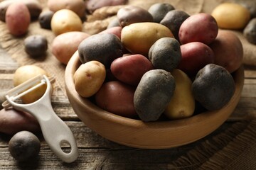 Different types of potatoes in bowl and peeler on wooden table, closeup