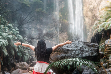Indonesian woman seen from behind raising arms in freedom while standing near a tropical waterfall. Concept of travel, adventure, happiness, nature connection, and spiritual relaxation.