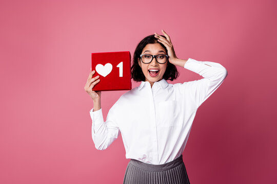 Young woman in white shirt holding a red like icon box, expressing excitement on a vibrant pink background