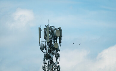 Cell tower with antennas against a blue sky with clouds and flying birds.
