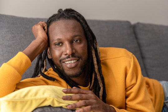 Smiling African american man with dreadlocks and yellow shirt relaxing on sofa at home - Powered by Adobe