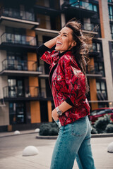 Stylish young woman smiling joyfully on a city street wearing a red printed jacket and jeans in daylight, vertical photo