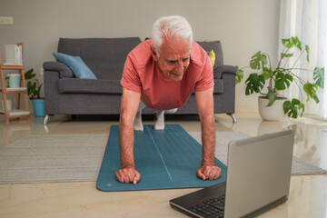 Senior man practicing push-ups on yoga mat following online fitness class on laptop at home