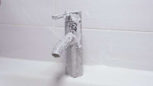 Gloved hand sprays descaler foam on limescale covered chrome faucet, hard water stains dissolve, bathroom maintenance restores shine.