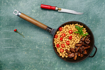 Savory grilled meat served with chickpeas, red chili pepper, and fresh herbs in a cast iron skillet. Set on a rustic green wooden table with ample copy space.