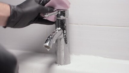Man in gloves cleans chrome bathroom faucet with foamy descaler, removes limescale and water spots, shared chores restore shine